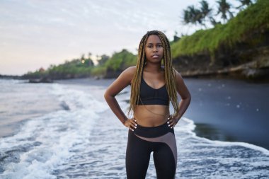 Koyu tenli ve okyanus ile spor sabah eğitimden sonra yıkanmış kıyısında duran güzel dreads ile motive genç kadın portresi. Kameraya bakarak eşofman giymiş Afro Amerikan kadın jogging yapan