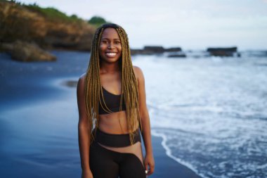 Okyanus su ile yıkanmış kıyısında ayakta dreads ile pozitif afro Amerikan sportwoman portresi. Deniz kıyısı kameraya gülümseyen iş için etkin kıyafetlerini giymiş genç kadın neşeli koyu derili