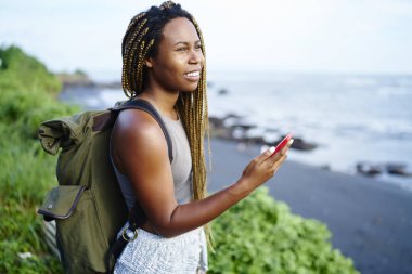 Neşeli cazip turistik koyu tenli smartphone elinde tutan sahil tropikal toplayan zevk. Manzara Adası gezisi sırasında hayran seyahat sırt çantası ile Afro Amerikalı genç kadın