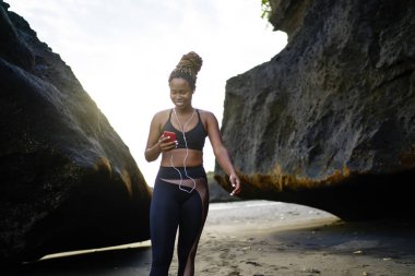 Pozitif afro Amerikan kadın jogger smarthone deniz kıyısı sabah eğitim için indirilen Player'da müzik seçimi etkin elbise giymiş. Neşeli genç kadın eşofman ve kulaklık