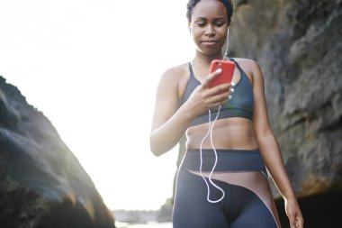 Genç kadın koyu tenli smartphone cep telefonu ile Player'da müzik şarkıları sabah egzersiz açık indiriyor. Afro Amerikan kadın runner etkin aşınmaya sahil eğitim gündoğumu sırasında için ses seçimi