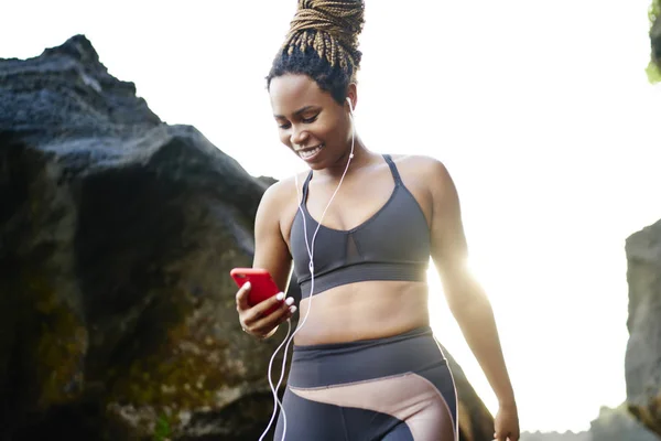 Cheerful afro american sportive woman in earphones connected to smartphone choosing music application for download on smartphone and morning training.Positive female in active wear and phone in hands