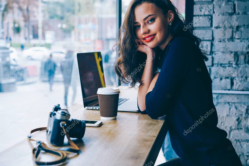 Portrait of attractive talented student sitting at modern laptop computer in coffee shop while looking at camera.Charming traveller enjoying leisure time in cafe with vintage camera and tasty beverage