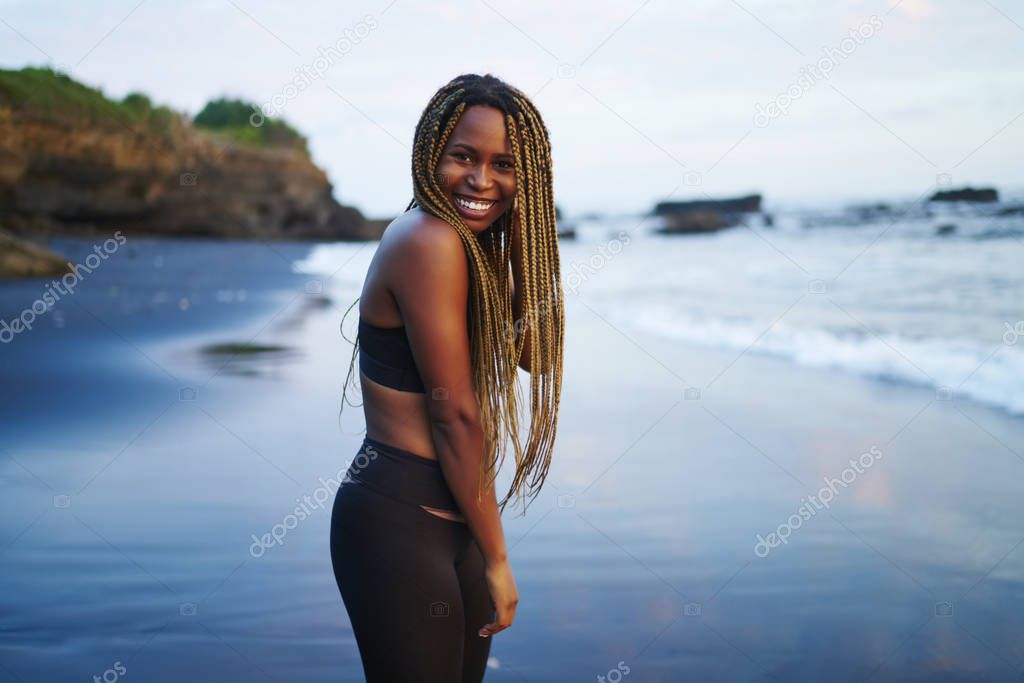 Portrait of cute afro american sportwoman with dreads smiling at camera.Cheerful young woman with dark skin dressed in stylish active wear standing on beautiful tropical beach washed by ocean water