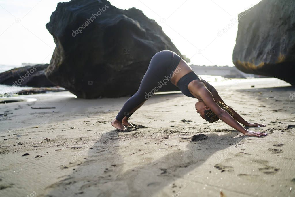 Afro american young woman dressed in active wear doing yoga exerciseses for healthy vitality on seashore of tropical island.Sportive girl with dark skin warming up before morning training on coastline
