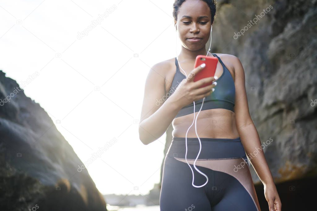 Young woman with dark skin downloading music songs in player on smartphone for morning workout outdoor.Afro american female runner in active wear choosing audio for training on seashore during sunrise