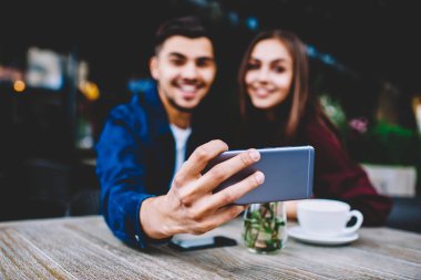 Seçici odak smartphone cep telefonu ile selfie için resim paylaşımı için cep telefonunuzun ağlarda yapma neşeli erkek ve dişi hipsters poz kafe terasında kız arkadaşıyla oturan genç adam elinde