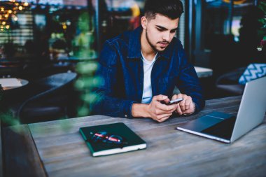 Düşünceli erkek öğrenci uygulamasında, dalgın hipster adam kafe iç çalışma üzerinde serbest uzak iş smartphone cep telefonu ile mesaj gönderme düzenleme için dizüstü bilgisayar ile telefon dosyaları eşitleme