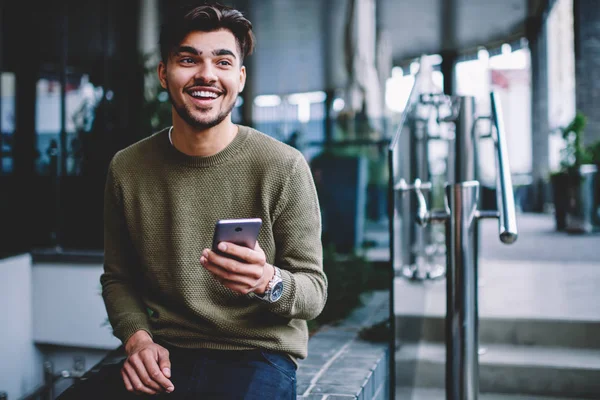 Happy bearded hipster guy excited with good news receiving message on smartphone sitting on urban settings, cheerful handsome man satisfies with mobile tariffs for calls and messages chatting on phone