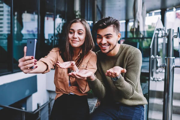 Happy male and female hipsters making funny photos for social networks posing on urban settings, cheerful best friends posing for smartphone camera making selfie while spending free time together