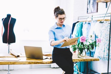 Şık gözlük profesyonel terzi kendi moda atelier dizüstü bilgisayar aygıt ve Tekstil giyim koleksiyonu için çizim kroki ile ahşap masaüstü üzerinde oturan çalışan yetenekli genç kadın