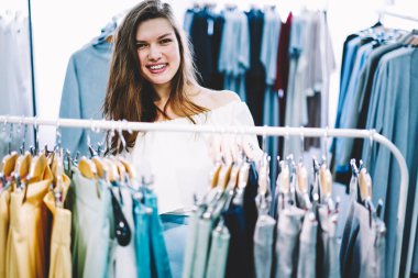 Kameraya bakarak ve raf arkasında trendy giyim duran Studio Tekstil giyim yeni koleksiyonu ile moda giysileri seçerken gülümseyen neşeli genç kadın müşteri yarım uzunluk portre