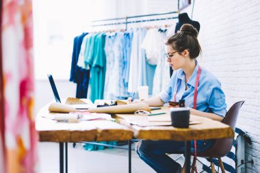 Yaratıcı kadın terzi gözlük ve ekipman ve modern latop bilgisayar kendi şık atelier içinde okul sırası vasıl oturan setches çizim yeni giysi tasarımı teyp yan görünüm