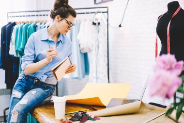 Masaüstünde oturma ve measuresin not defteri denetimi sarı kroki bakarak gözlük şık showroom konsantre kadın sahibi. Dalgın terzi malzemeleri elbise oluşturmak için hazırlanıyor