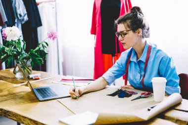 Gözlük ve teyp modern latop bilgisayar kendi atelier Studio ile dikiş masaüstünde oturan şık giysiler için yeni tasarım kroki çizim önlemler için konsantre profesyonel tasarımcı