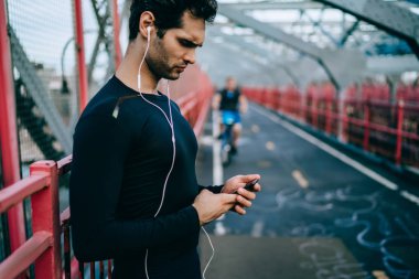 Yan görünüm sporcu eğitim müzik uygulama smartphone cep telefonu ile ses şarkıları seçmek için eşofman giymiş. Yakışıklı atletik delikanlı oyuncu kentsel ortamda ayakta dinlerken kulaklık