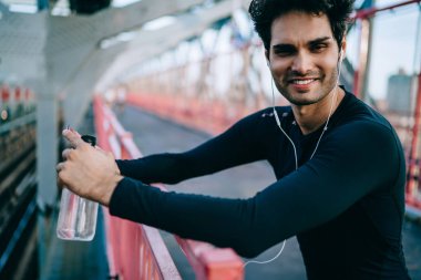 Neşeli erkek atlet portresi kameraya gülümseyen kulaklık ses dinleme siyah aktif giyim giymiş. Eşofman ayakta şişe elinde tutan kentsel ortamda pozitif sporcu