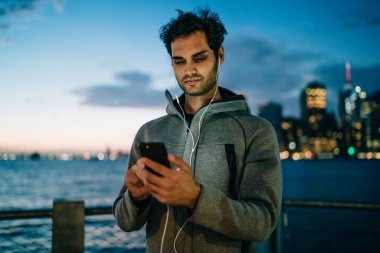 Online sosyal ağlar arkadaşlar ve dinleme radyo earphones ile mesajlaşma yakışıklı genç adam akıllı telefon açık havada akşam saat metropolis arka plan üzerinde ayakta bağlı