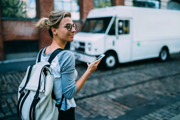 Smiling female tourist walking on city street checking route for navigate via application on smartphone,positive young hipster girl with backpack using mobile phone for calling taxi and paying online