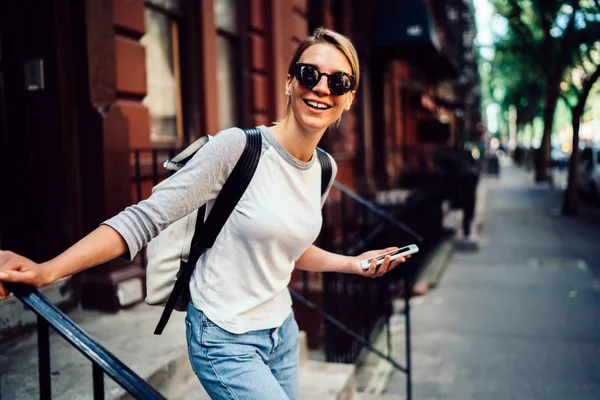 Half length portrait of cheerful young woman leaving building laughing holding telephone,smiling hipster girl in sunglasses spending weekends actively in good mood strolling in downtown with smartphon