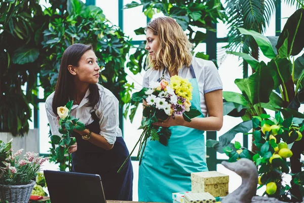 Female colleagues working as florist in botanic orangery cooperating during creating floral composition,young woman in apron assist professional owner of flower store making bouquet for clients orde