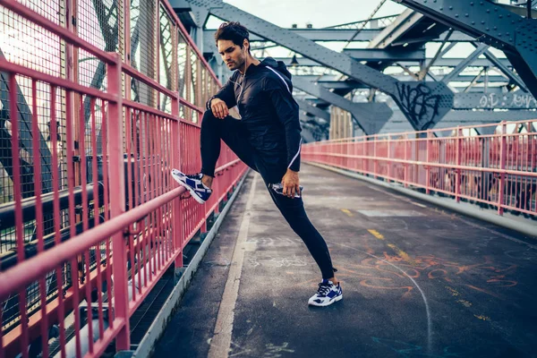 Young man in active wear listening music in earphones and warming up before training on modern bridge.Sportive man in black tracksuit and sport sneakers having workout outdoors while enjoying songs