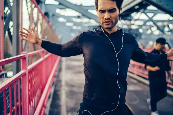 Bearded fit man in active clothes stretching hands during warming up on birdge while listening music player in earphones.Sportsman dressed in tracksuit in headphones doing sport exercises outdoors