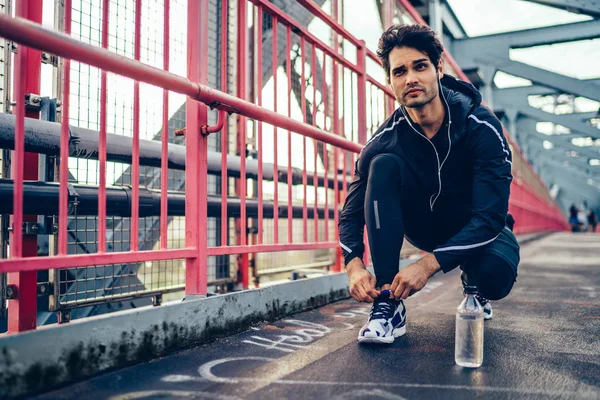 Professional sportsman dressed in active tracksuit listening music player in earphones and tied up laces on stylish sneakers during morning workout in urban setting on bridge for getting sportive goal