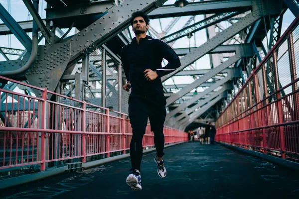 Motivated male jogger doing cardio to maintain sporting form in urban setting.Young athlete dressed in black tracksuit running on bridge.Fit man in active wear and sneakers training outdoors