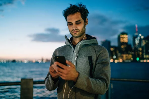 Online sosyal ağlar arkadaşlar ve dinleme radyo earphones ile mesajlaşma yakışıklı genç adam akıllı telefon açık havada akşam saat metropolis arka plan üzerinde ayakta bağlı