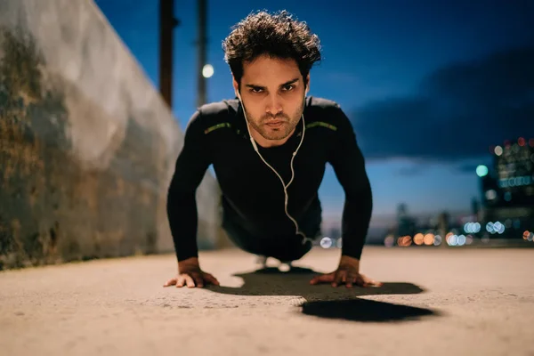 Portrait of motivated fit man in active wear listening music in earphones and pushing up from ground in twilight.Serious sportsman doing sport exercises during evening workout to maintain vitality