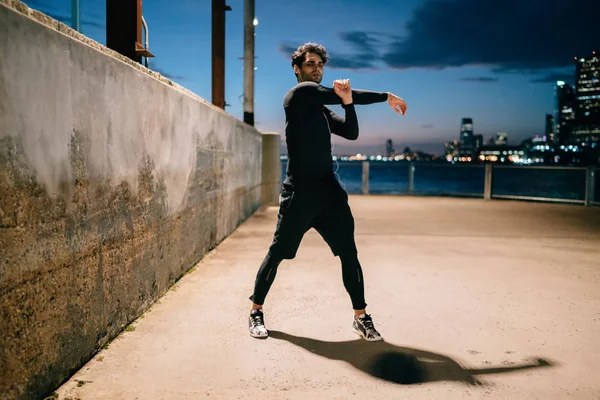 Portrait of professional male jogger dressed in sport wear doing stretching of hands training outdoors in evening time.Sportsman in tracksuit and sneakers warming up before run in urban setting at night