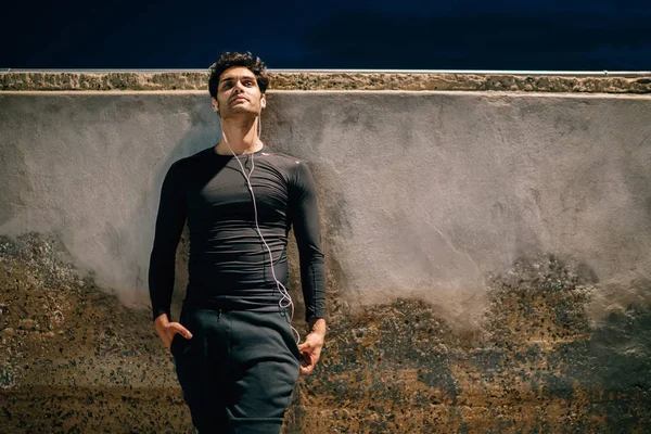 Thoughtful sportsman dressed in active wear for training holding hands in pocket and listening music from playlist in earphones while standing opposite wall with copy space for your sport advertising