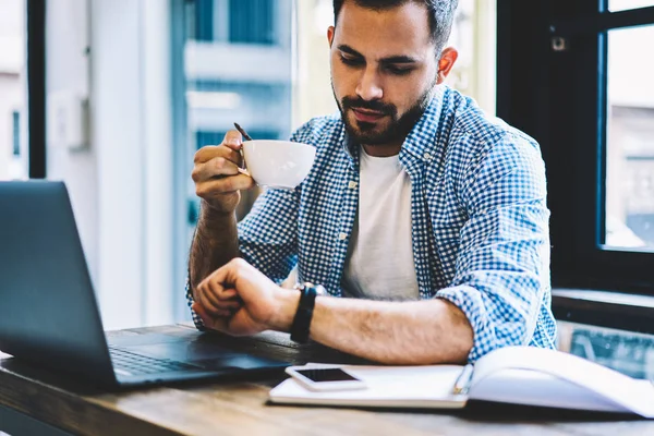 年轻的高加索男性 开发人员看手表在咖啡休息时坐在 Coworking 办公室与上网本 学生检查时间持有杯等待笔记本电脑上的软件更新 — 图库照片