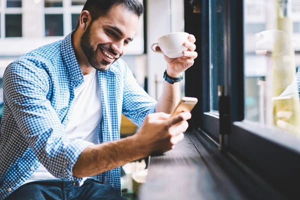 Cheerful male person happy to receive messages from friend checking mail via smartphone during rest in cafe, smiling man satisfied with getting good news using telephone fr networking drinking coffee