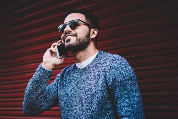 Young hipster guy smiling while talking on mobile phone using cheap tarifs in roaming standing on urban settings,male person in trendy outfit having telephone conversation satisfied with connection