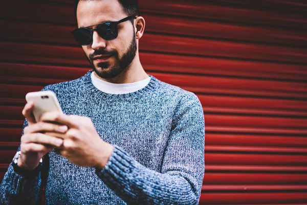 Handsome young man sending text message chatting on smartphone standing on urban settings background,hipster guy checking notification on cellular updating application spending free time outdoors