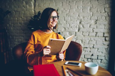Şık coworking uzayda oturan bestseller ilginç komik çizim zevk elinde mutlu güzel okuyucu kitap ile. Literatür bilgileri öğrenme sırasında gülüyor gözlük olumlu hippi kız