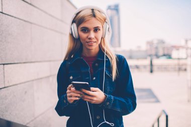 Modern Kulaklığımda yabancı dil öğrenme dinleme ses ders smartphone aygıt açık havada ayakta bağlıyken kameraya arayan çekici sarışın öğrenci portresi yarım uzunluk