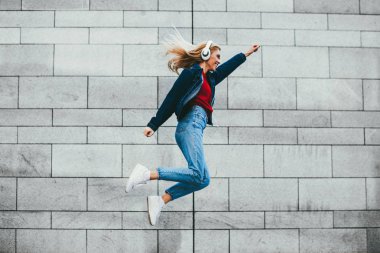 Neşeli genç kadın açık havada gri duvara atlama rahat denim giyim giymiş. Pozitif hippi kız kot pantolon headpones promosyon arka planda çalma listesinden müzik dinleme serin giysiler