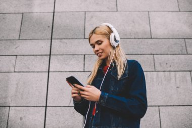 Modern Kulaklığımda çalma listesinden serin müzik dinleme sırasında e-posta kontrol denim ceket gülümseyen hippi kız Smartphone-e doğru bağlı. Neşeli erkek sesli telefon açık havada yürüyüş üzerinde kitap İndiriyor