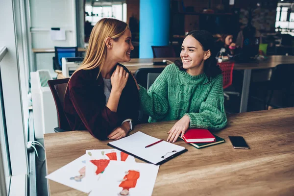 Two best friends laughing during break sitting at desktop with design sketches for new fashion collection in studio interior.Young women communicating with each other about successful project