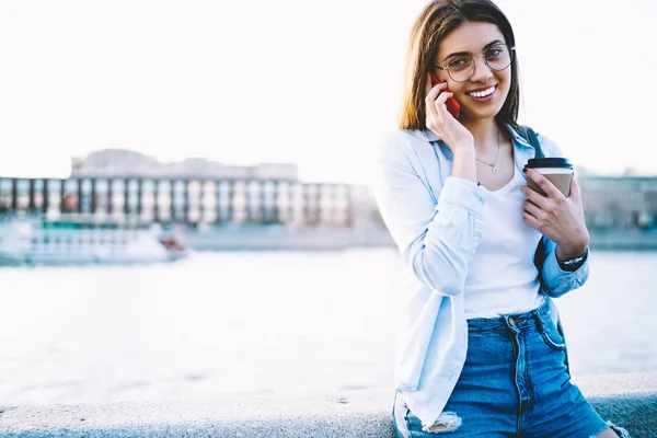 Portrait of cheerful hipster girl with coffee to go talking on mobile phone standing near copy space for advertising outdoors, smiling female traveler using roaming for making international calls