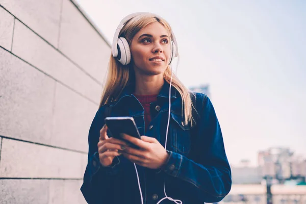 Charming young woman in denim outfit choosing audio songs to download on mobile player via 4G internet connection.Blonde hister girl in modern headphones listening music standing on street