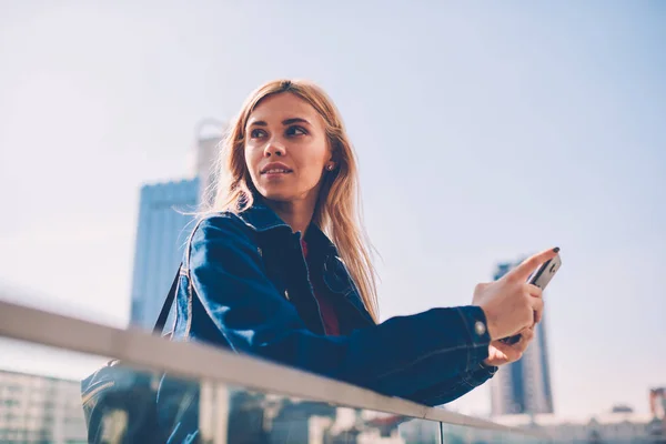 Attractive blonde young woman in denim jacket looking away while installing app on smartphone standing on street.Charming female tourist updating profile in social networks on phone via internet