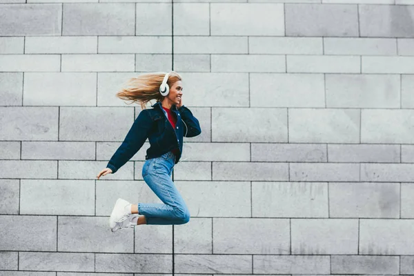 Carefree young woman with blonde hair listening favourite electronic music from playlist in modern white headphones in the air during jump on grey promotional background enjoying good mood