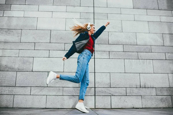 Side view of cheerful female meloman dressed in denim apparel listening audio records from playlist in modern headphones during jumping jacks outdoors on grey promotional background with copy space