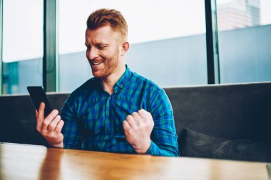 Mutlu genç adam web sitesinde online oylama kazandı. Memnun hipster adam Kızıl saçlı Internet ikramiye ve smartphone aygıt üzerinde alınan diğer tarif planları için bedava dakika sms mesajı okuma