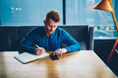 Dalgın zeki öğrenci bilgi coworking içinde oturan smartphone cep telefonu ile Internet bağlantısı kullanarak Web sitesinden yeniden yazma. Uzaktan eğitim Not Defteri'nde telefon kayıtlarından belirterek Kızıl saçlı genç adam