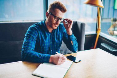 Gözlük aşağı boşaltmak boşluk coworking içinde ahşap masa öğrenim notları yazma akıllı genç adam. Planlama çalışma süreci kafede oturan gözlük zeki hipster adam